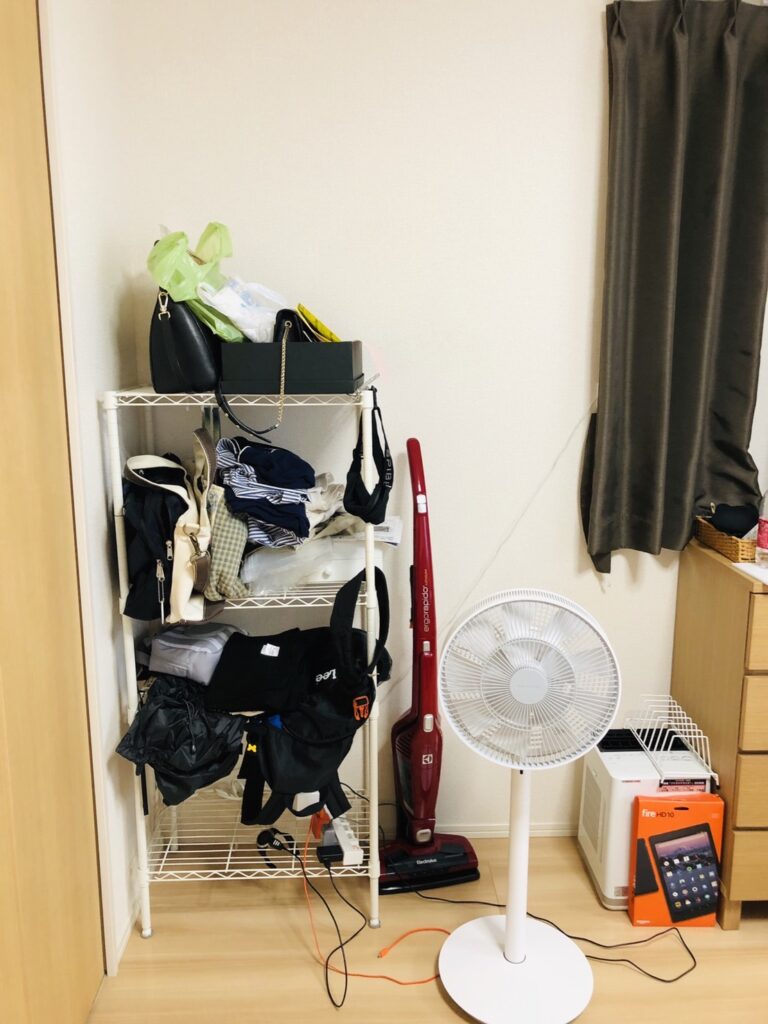 バッグや衣類が雑然と重なったワイヤーラックと掃除機。断捨離前の汚部屋の様子